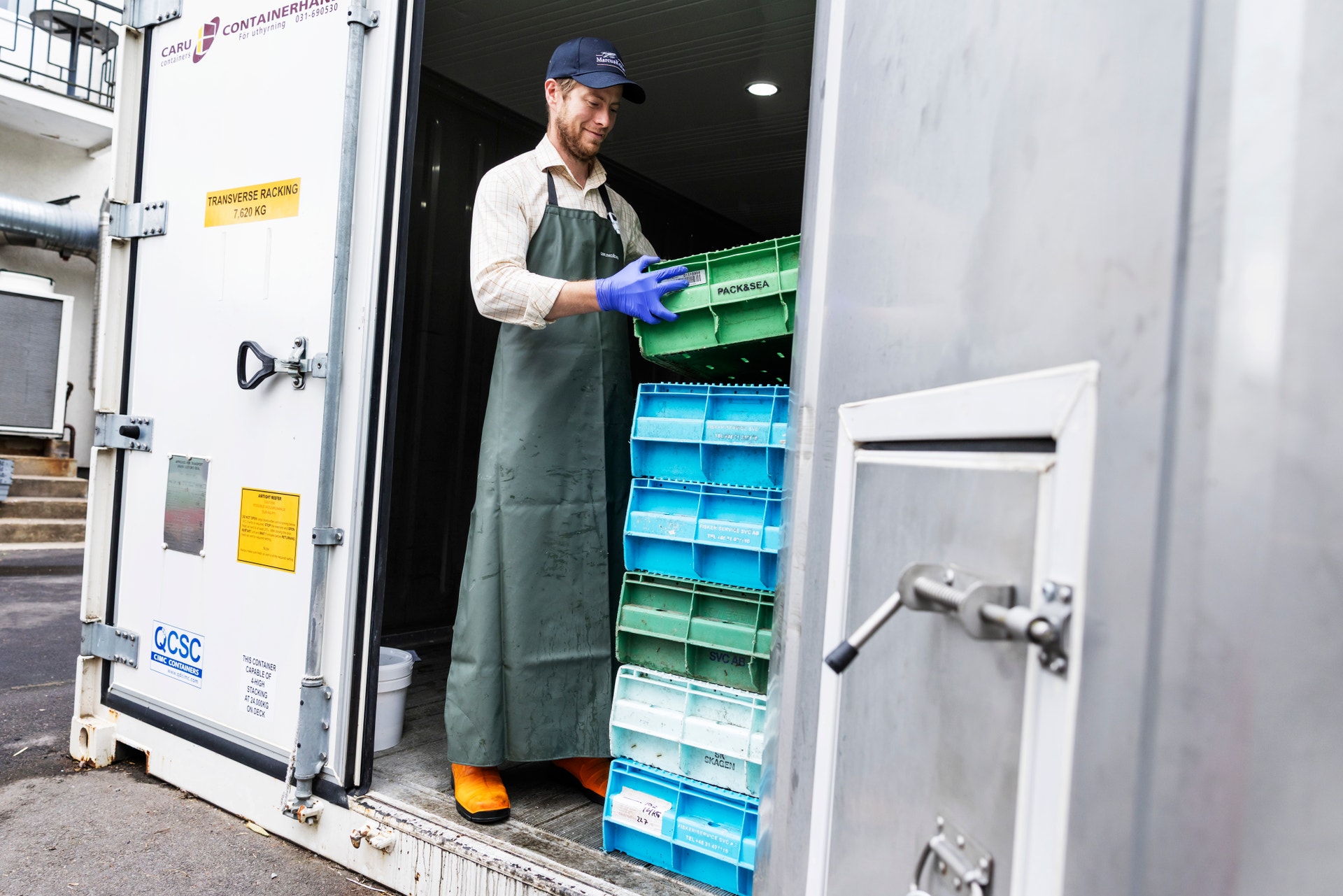 En man i arbetskläder och blå handskar hanterar färgade plastlådor i en vit container.