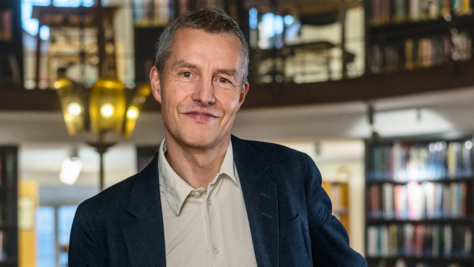 Porträtt av en man i mörkblå kavaj och ljus skjorta framför en bokhylla i en modern inomhusmiljö.