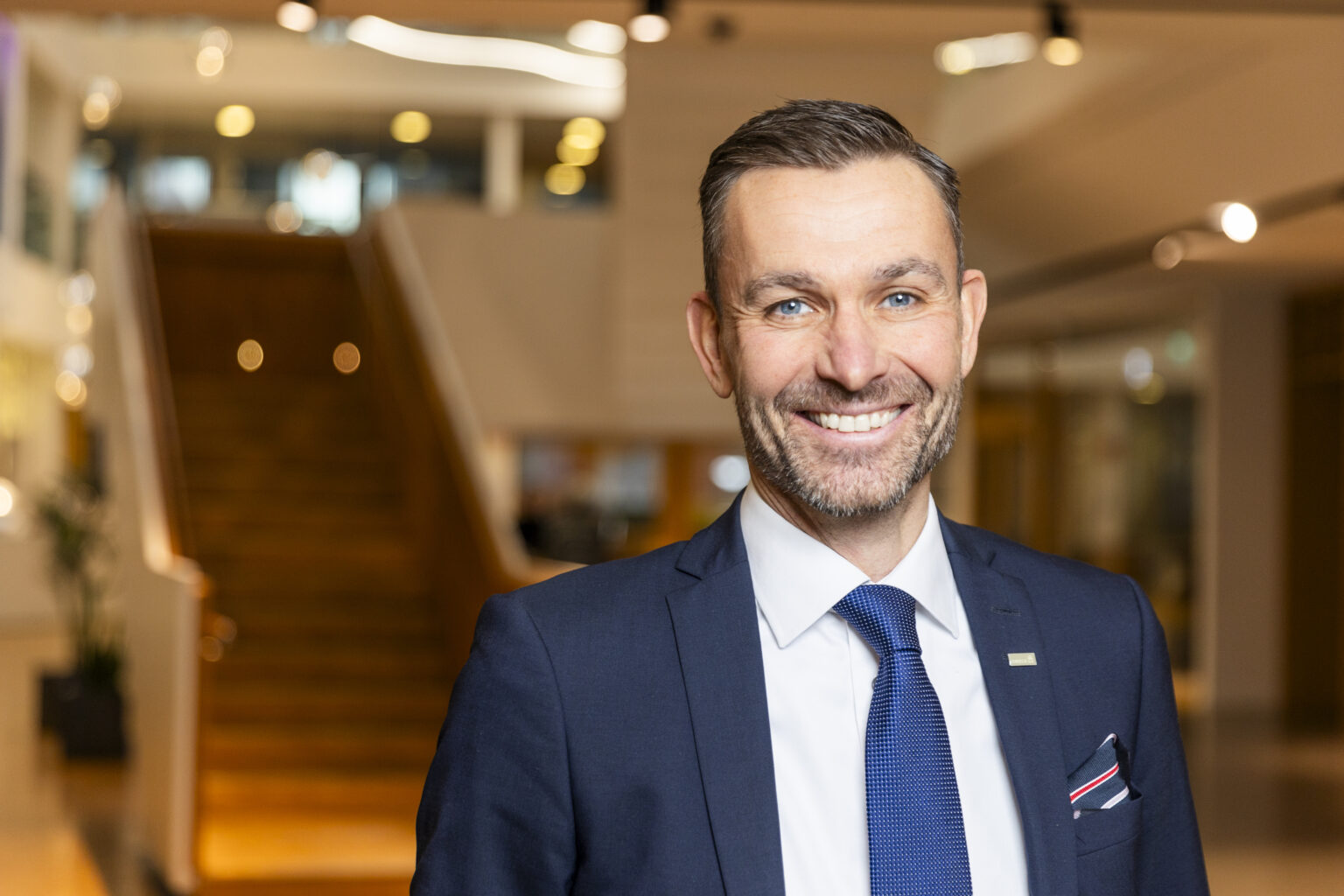 En leende man i kostym och slips står i en ljus entréhall med en trappa i bakgrunden.