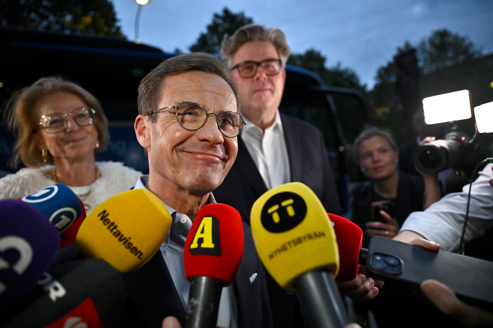 En leende man intervjuas utomhus av flera journalister som håller fram mikrofoner mot honom.