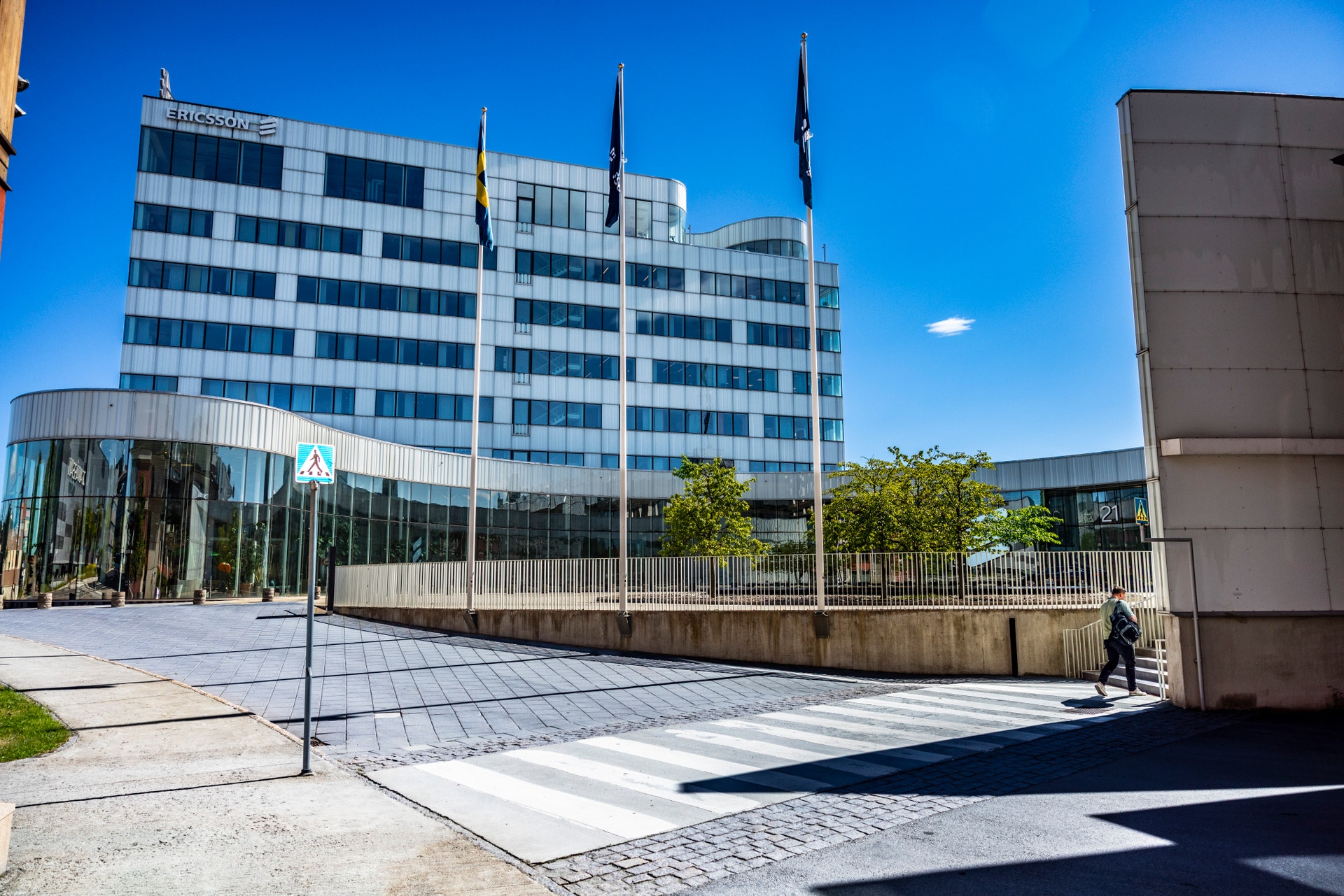 Modern kontorsbyggnad i Kista med vita och blå glasfasader, flaggor på taket, omgiven av gröna träd och en öppen plats framför byggnaden under blå himmel.