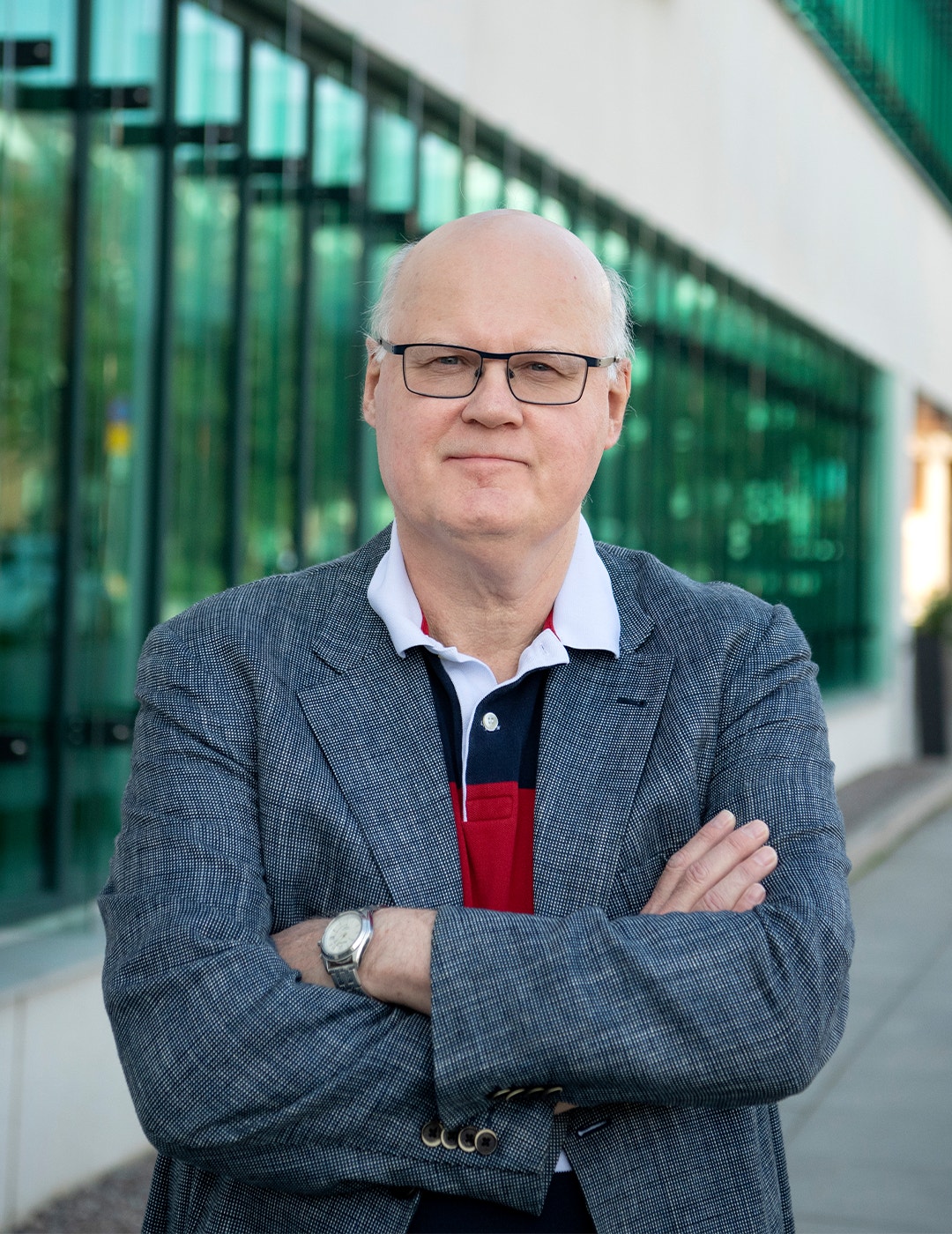Porträtt av en man med glasögon som står framför en modern byggnad med gröna glasfönster. Han bär en grå blazer och har armarna i kors.