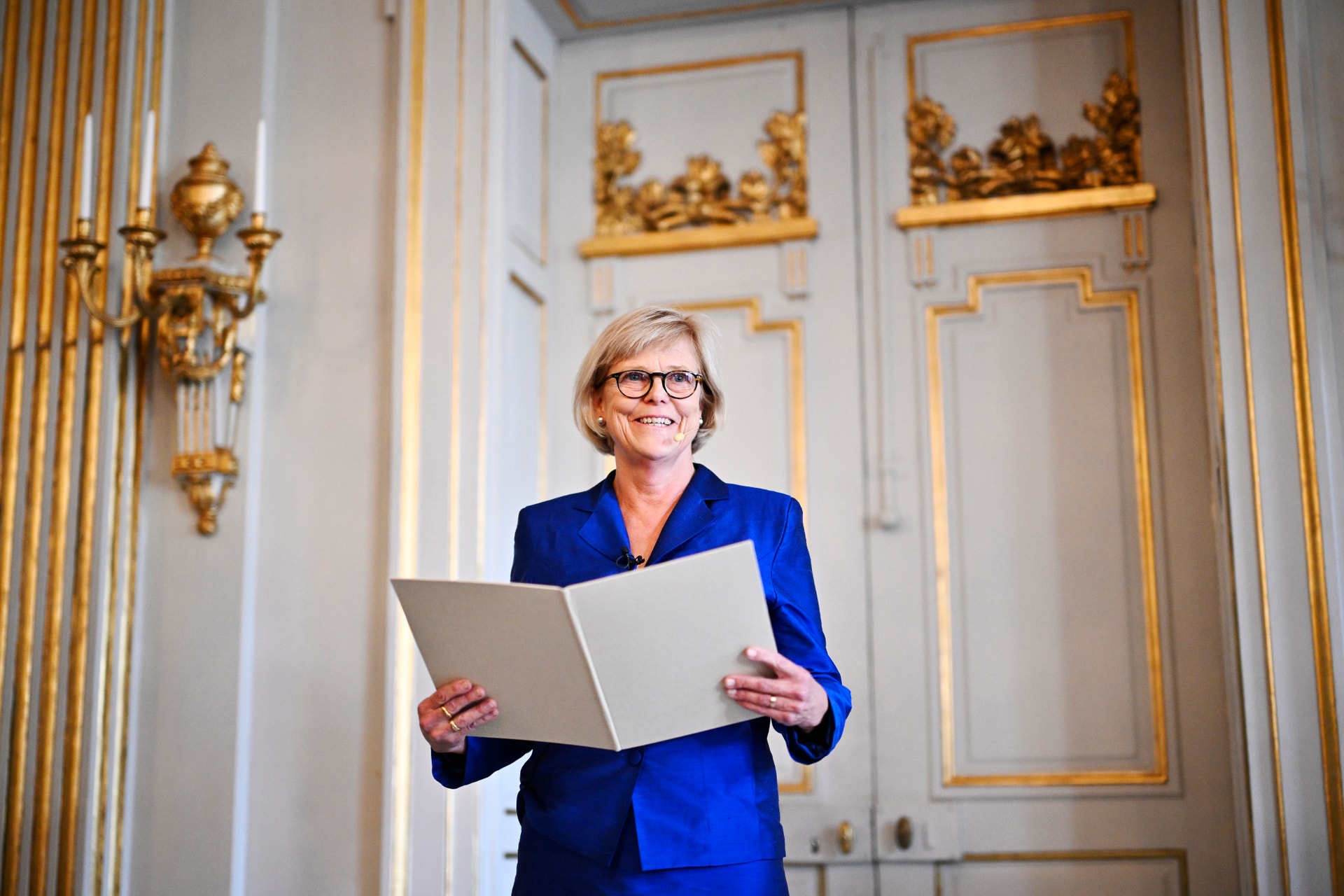 Leende kvinna i blå kavaj står med ett uppslaget dokument i händerna i en ljus sal med gulddetaljer.