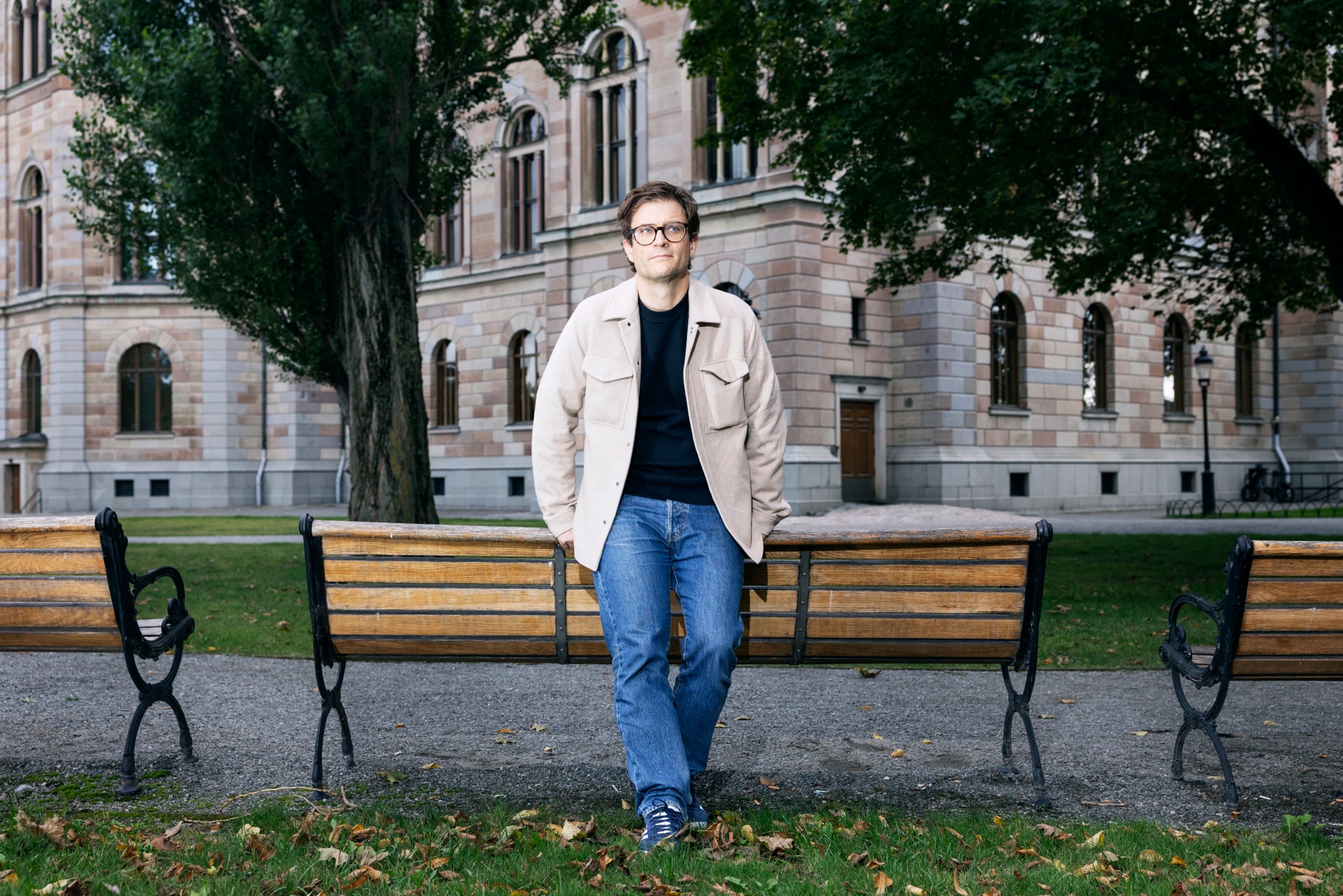 Porträtt av en person som står mellan två träbänkar på en gräsplan framför en klassisk stenbygnad.