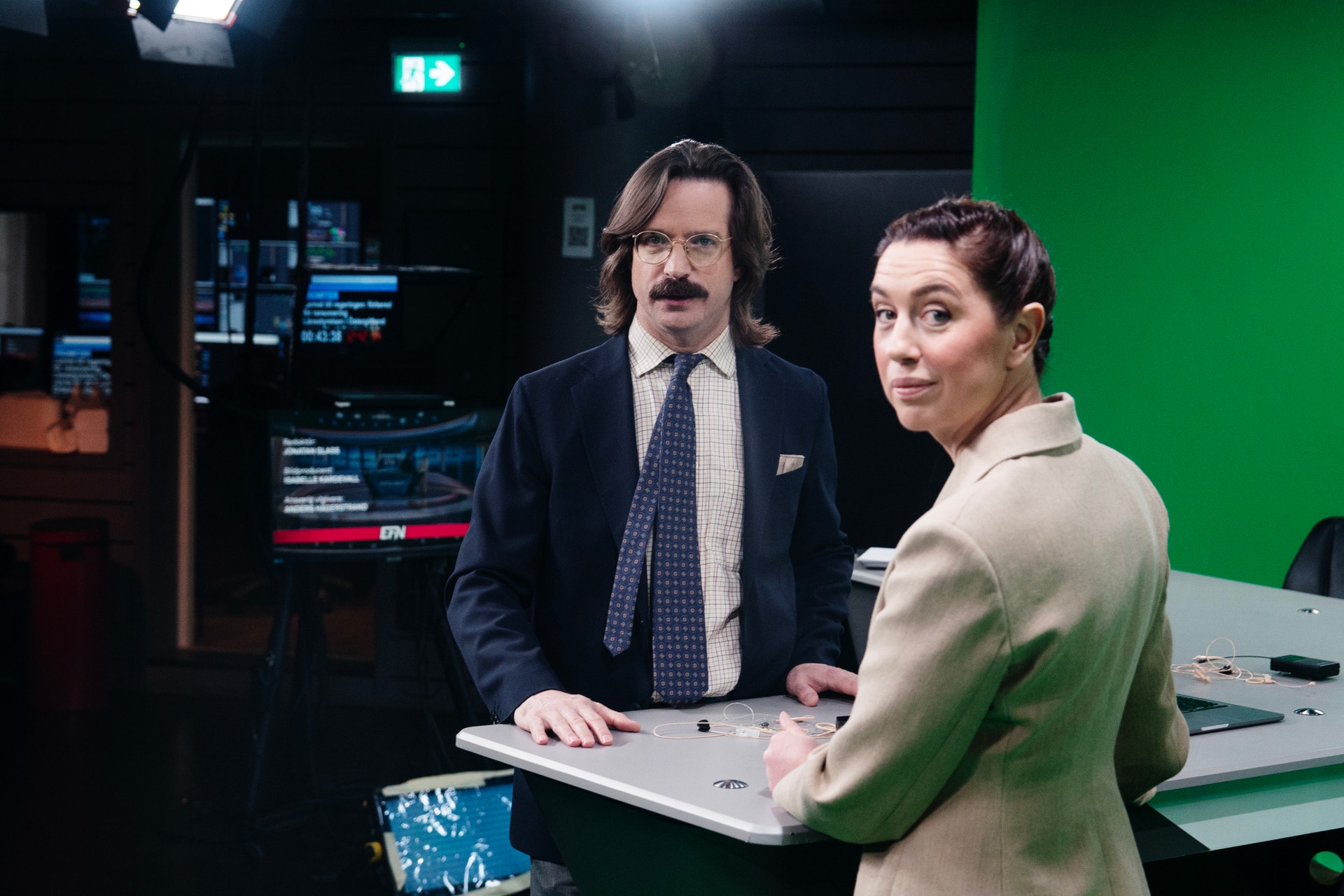 En man och en kvinna står vid ett bord i en studiomiljö med teknisk utrustning och grön skärm i bakgrunden.