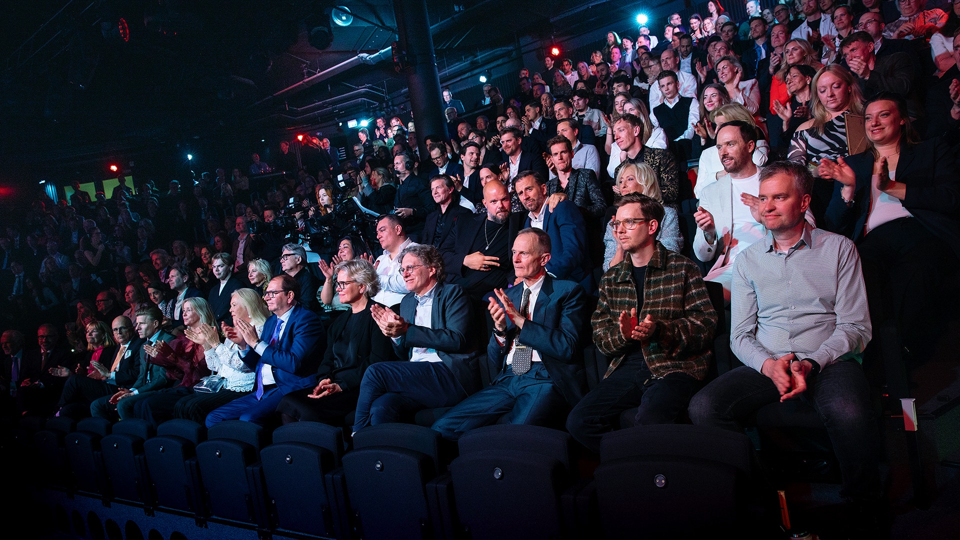 En stor publik sitter och står i en mörk sal under ett event. Människorna är upplysta av blått och rött scenljus och verkar engagerade och uppmärksamma.