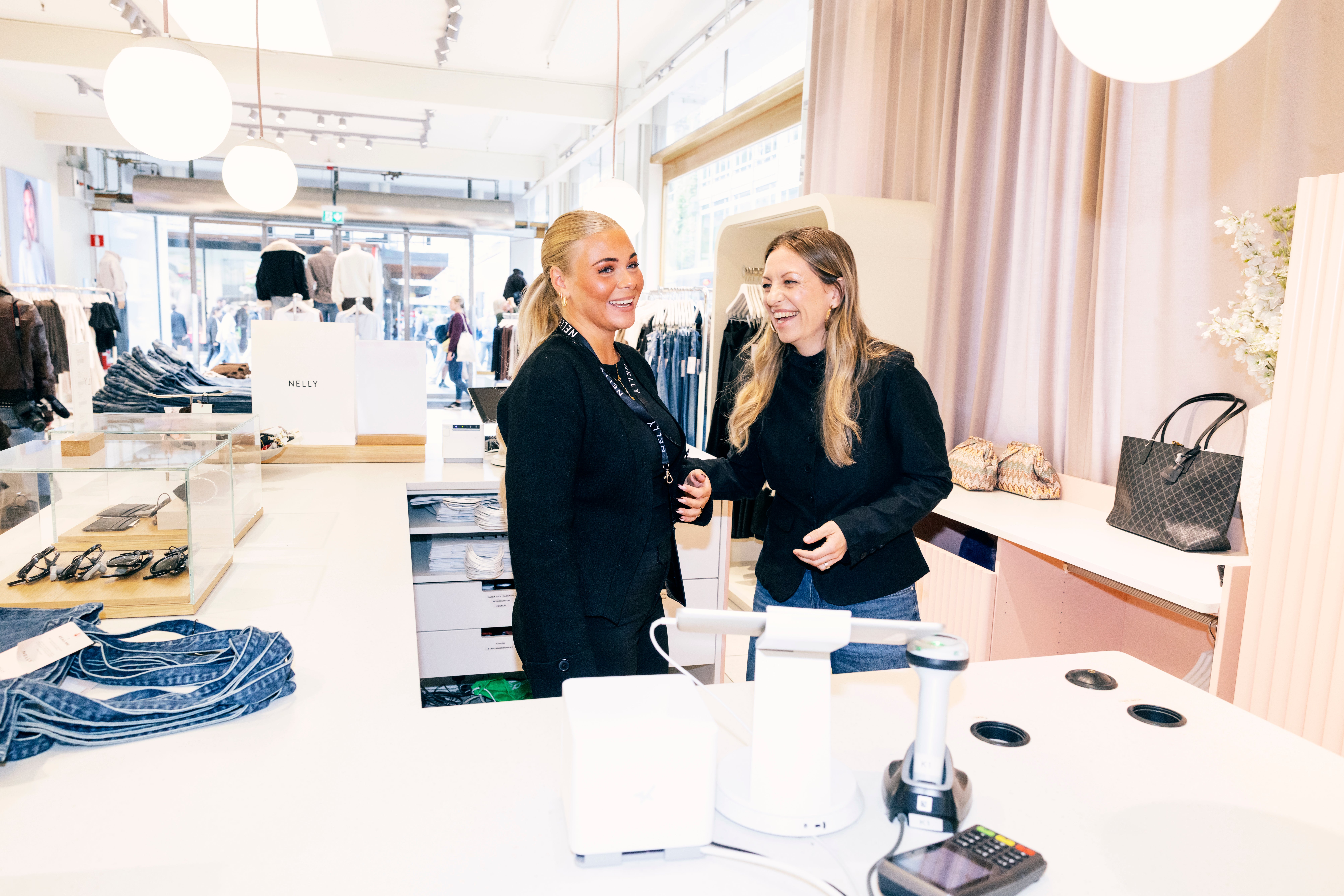 Två kvinnor i svarta kläder står vid en vit disk i en modern butik och samtalar. I bakgrunden syns kläder och accessorier på display.
