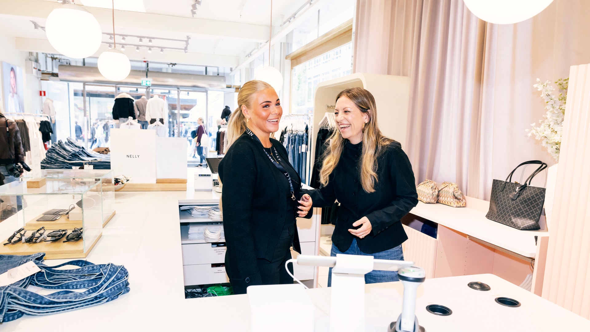 Två kvinnor i svarta kläder står vid en vit disk i en modern butik och samtalar. I bakgrunden syns kläder och accessorier på display.