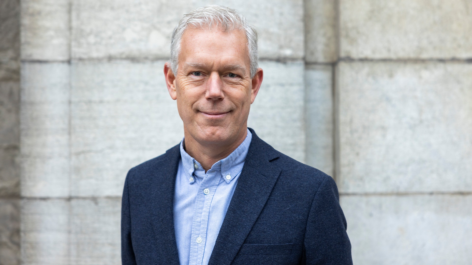 Porträtt av en man i mörkblå kavaj och ljusblå skjorta framför en stenmur.