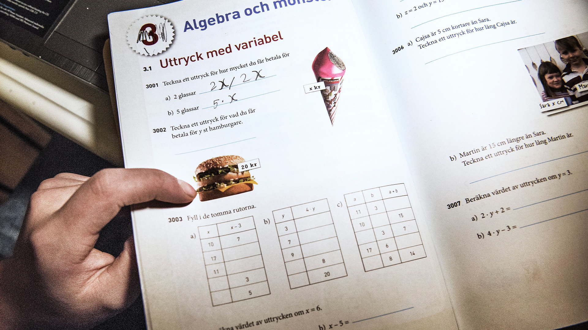 En person håller i en hamburgare medan hen läser ett matematikpapper med algebrauppgifter.