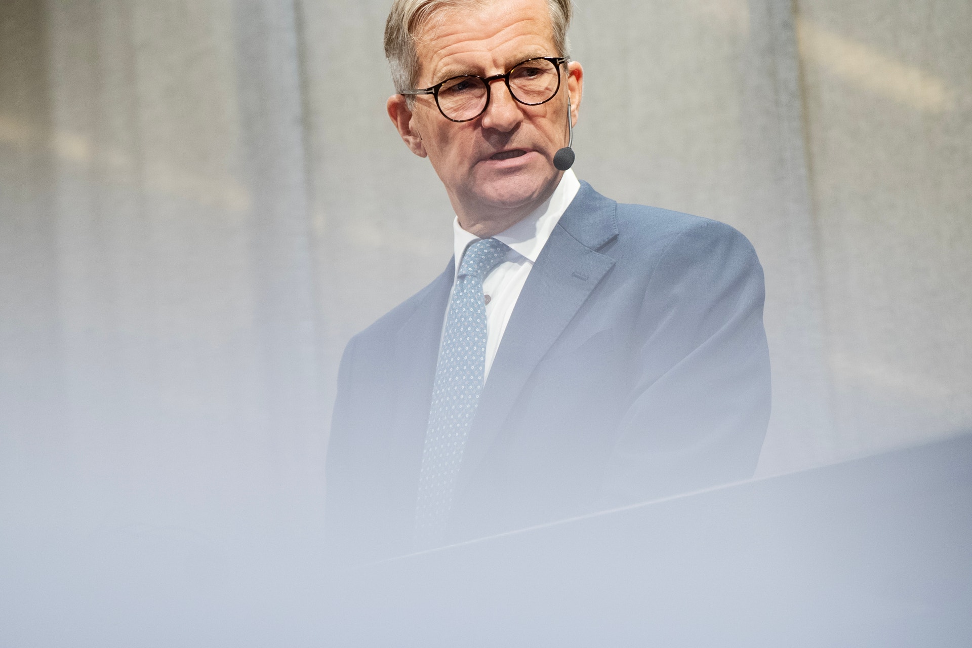 Porträtt av en man med glasögon, blå kostym och slips mot ljus bakgrund.