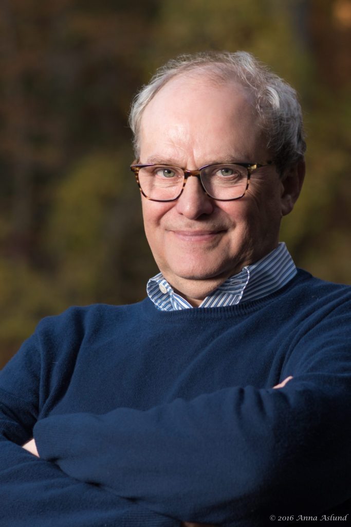 Porträtt av en äldre man med glasögon, klädd i blå tröja, som ler mot kameran utomhus.