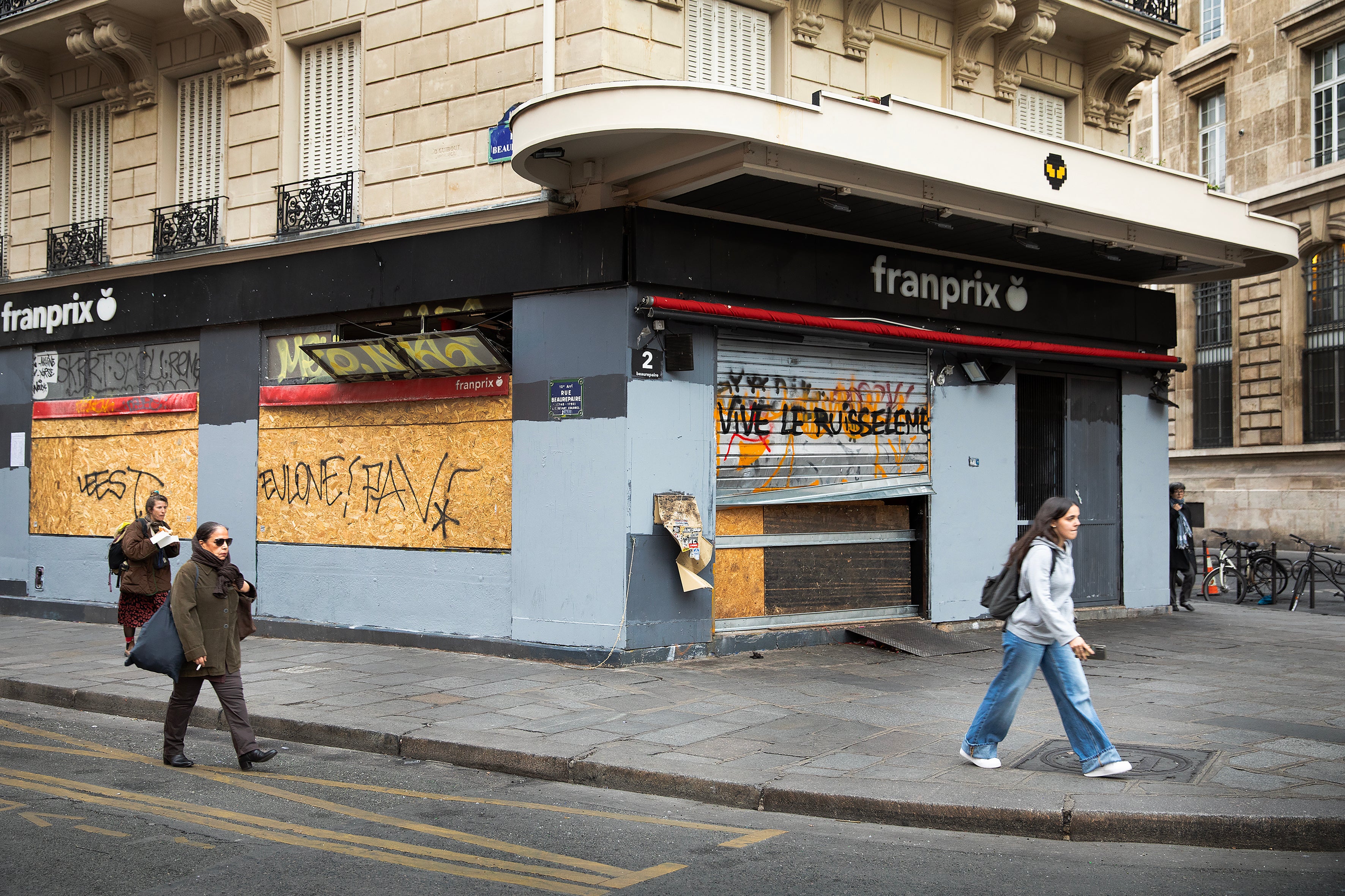 Stängd och barrikaderad livsmedelsbutik på ett gathörn i Paris. Fönstren är täckta med träskivor och graffiti, medan några personer går förbi på trottoaren.