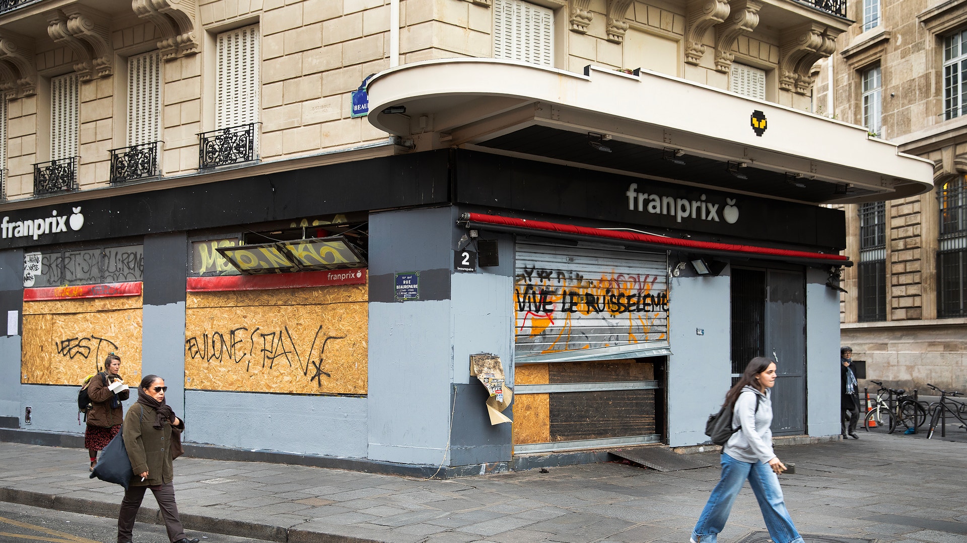 Stängd och barrikaderad livsmedelsbutik på ett gathörn i Paris. Fönstren är täckta med träskivor och graffiti, medan några personer går förbi på trottoaren.