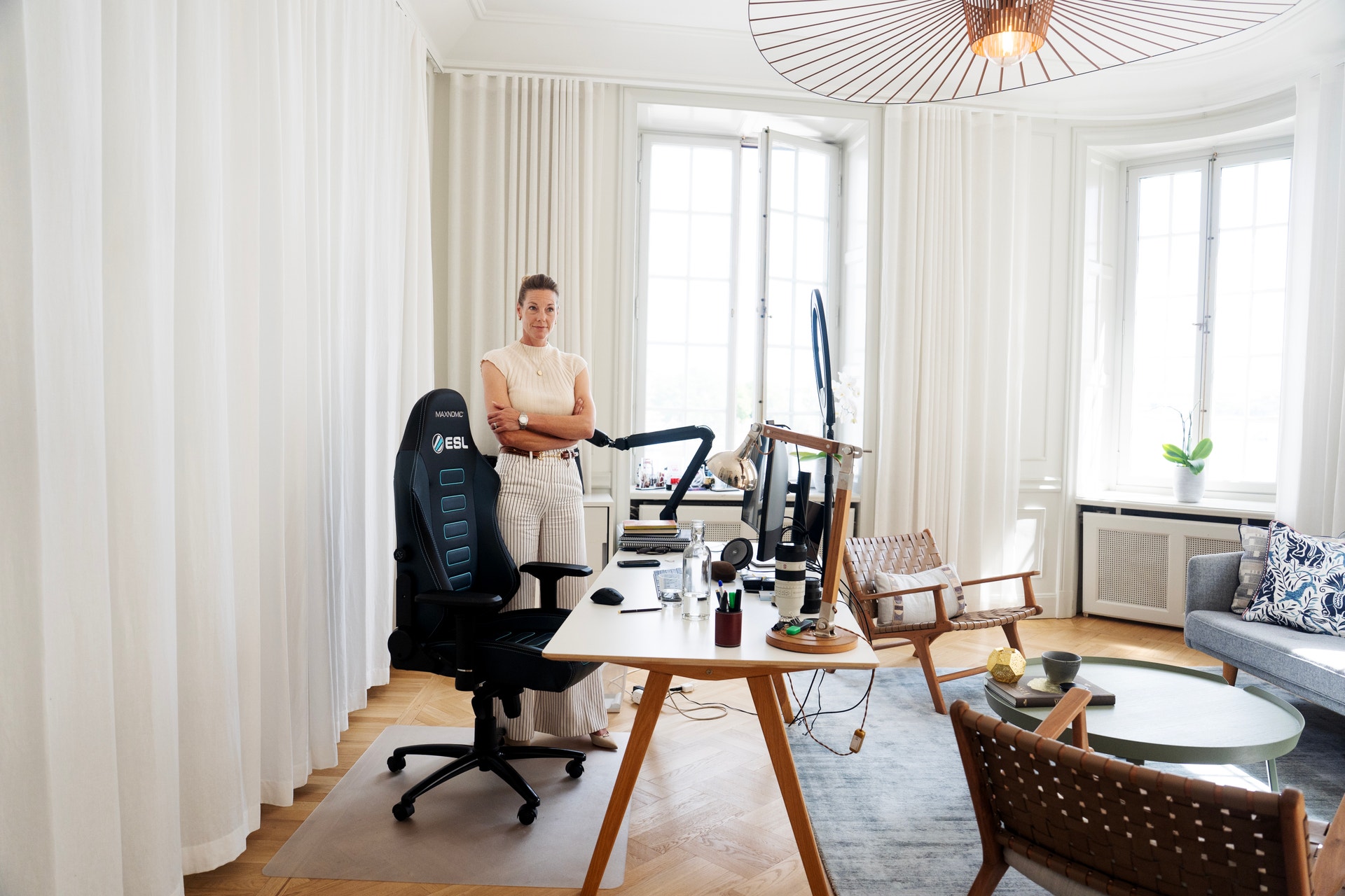 En kvinna står vid sitt skrivbord i ett ljust hemmakontor med stora fönster och vita gardiner.