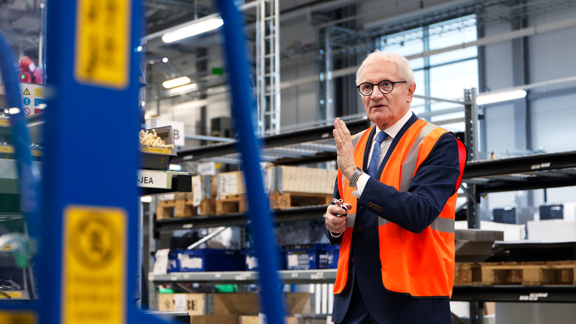 Man i orange säkerhetväst och glasögon står i ett lager och inspektera eller presenterar något. Blå och gula metallhyllor med lådor syns i bakgrunden.