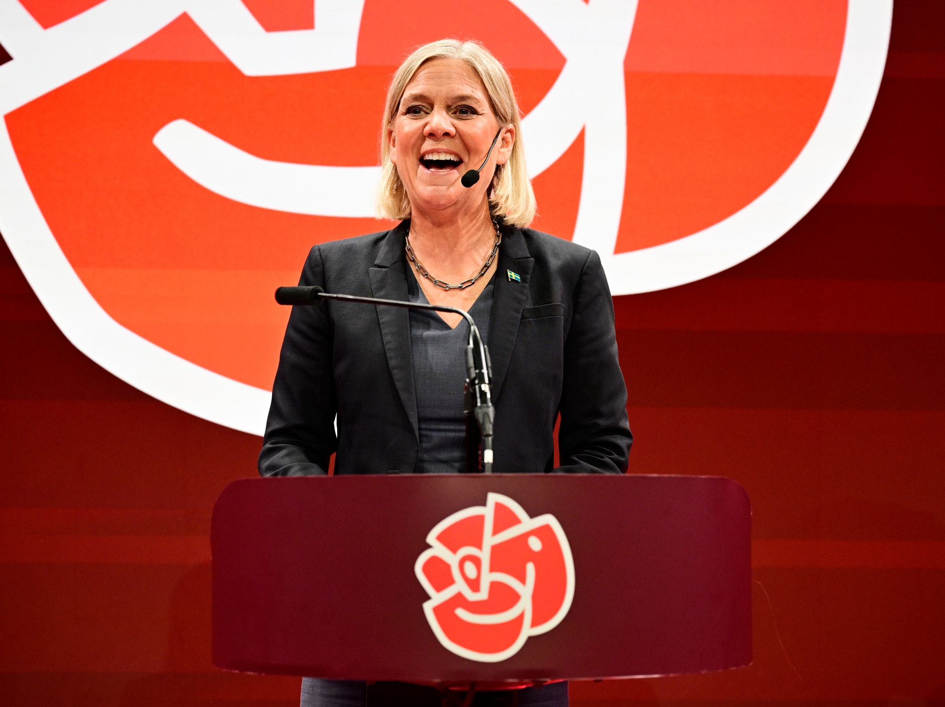 En leende kvinna talar i mikrofon bakom en talarstol med Socialdemokraternas logotyp.