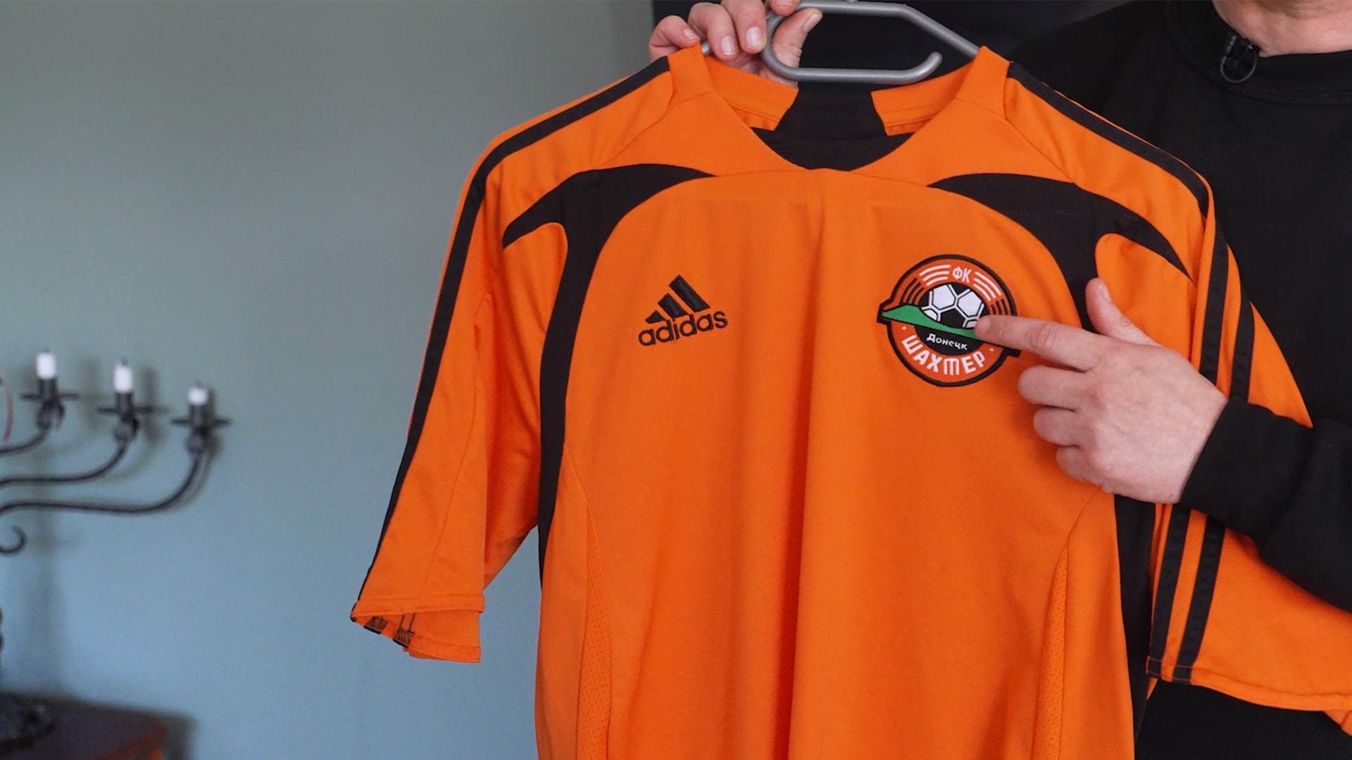 Orange Adidas-tröja med svarta detaljer och ett klubbmärke på bröstet, hållen upp av en person.