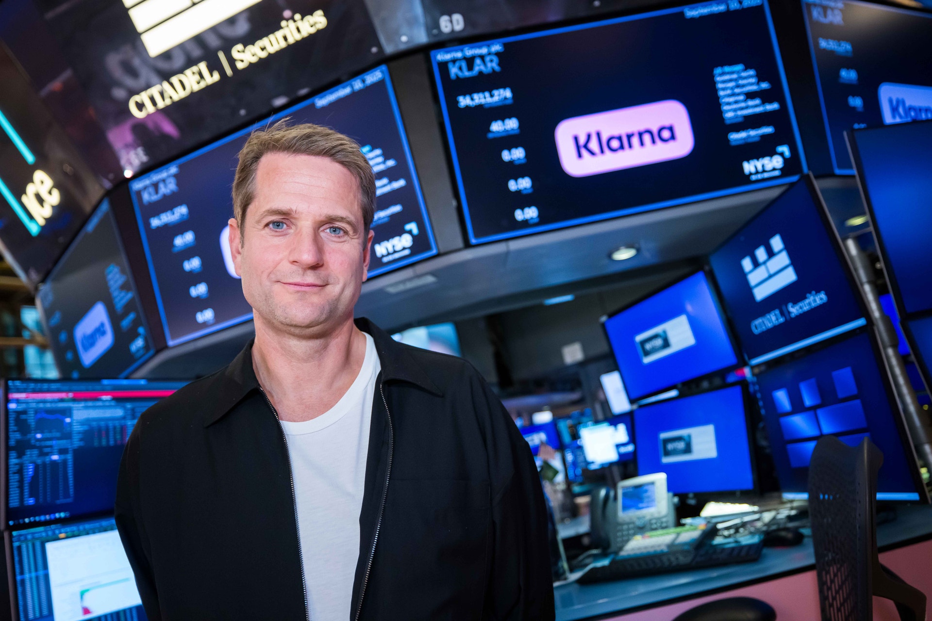 En man står framför skärmar som visar börsinformation och Klarnas logotyp.