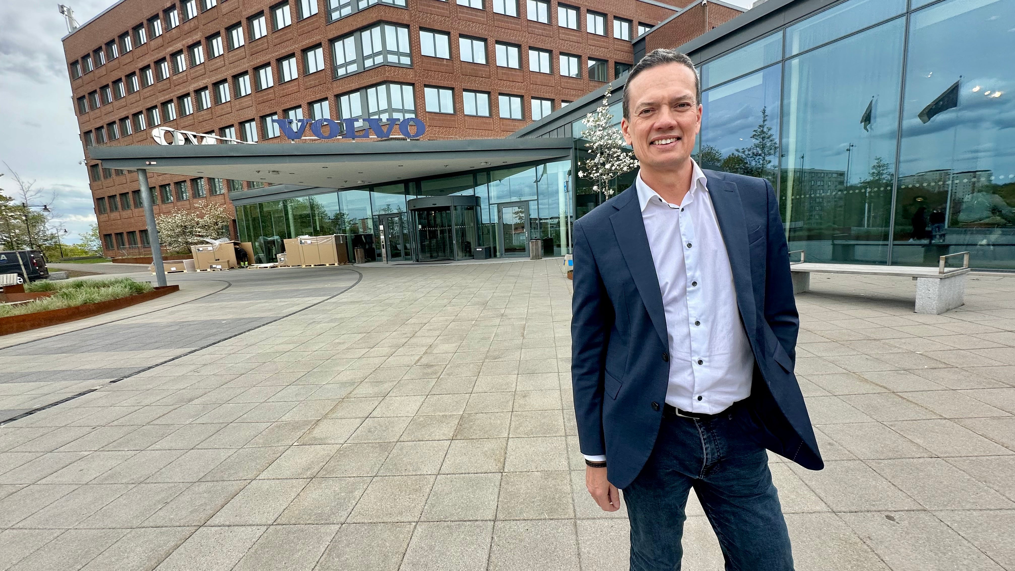 En man i mörkblå kavaj och vit skjorta står på en stenlagd plats framför moderna byggnader.