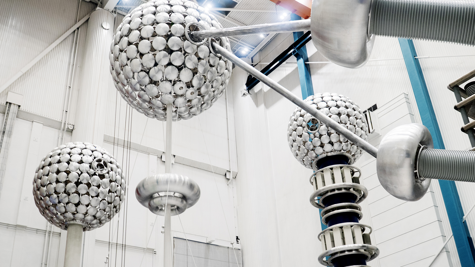 Industriell högspänningsutrustning med sfäriska elektrodkupoler monterade på metalliska stöd i ett testlaboratorium. Utrustningen består av två stora vita kulformade elektroder och en mindre elektrod på höger sida, anslutna med metalliska ledningar och isolatorer för högspänningstest.