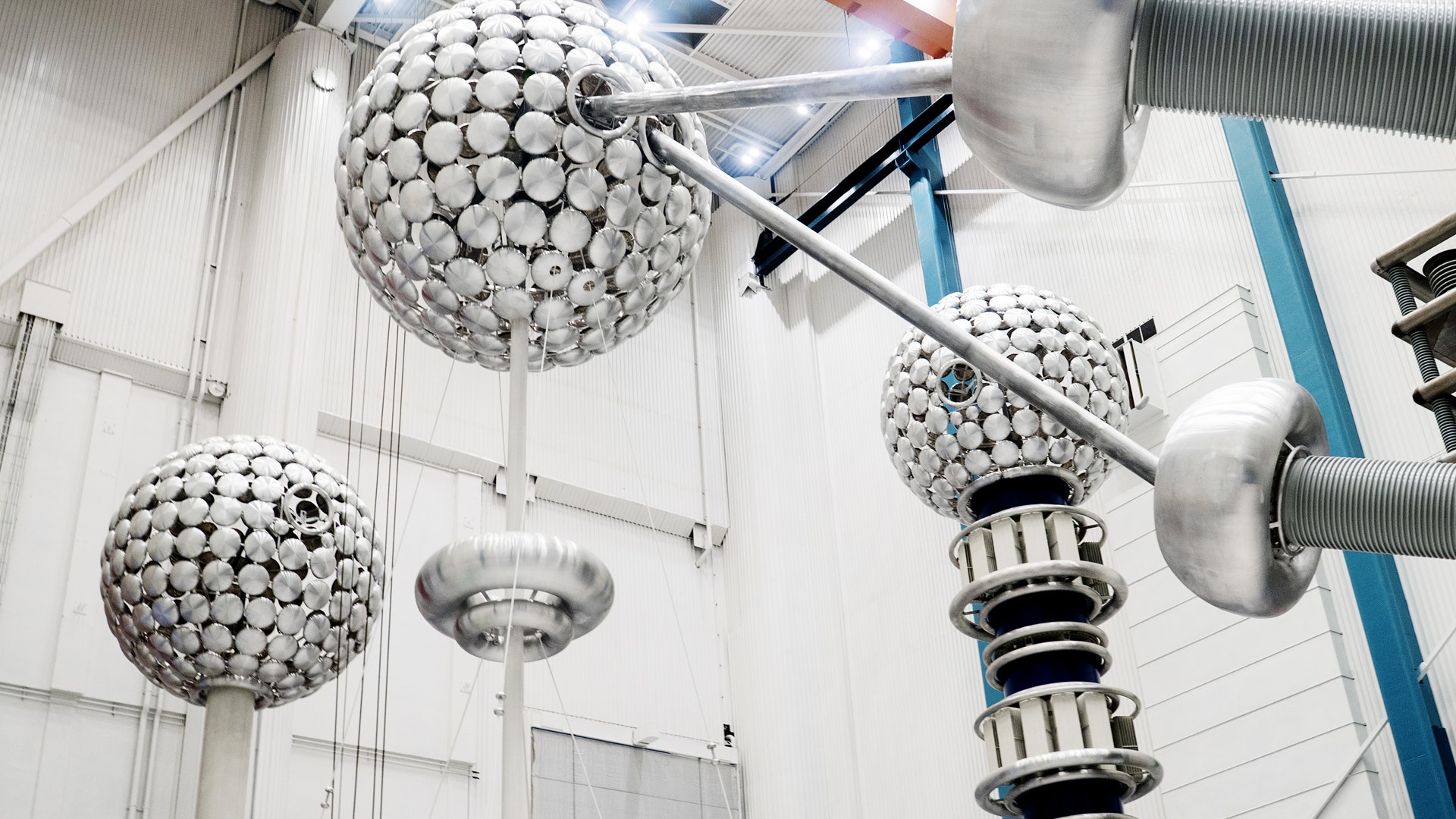 Industriell högspänningsutrustning med sfäriska elektrodkupoler monterade på metalliska stöd i ett testlaboratorium. Utrustningen består av två stora vita kulformade elektroder och en mindre elektrod på höger sida, anslutna med metalliska ledningar och isolatorer för högspänningstest.