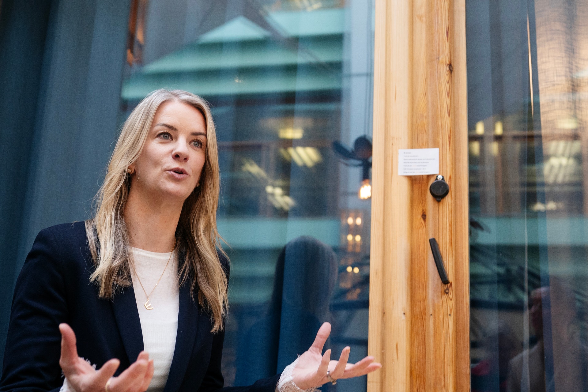 Kvinna i svart kavaj och vit skjorta talar framför en glaskonstruktion med stadsmiljö i bakgrunden.