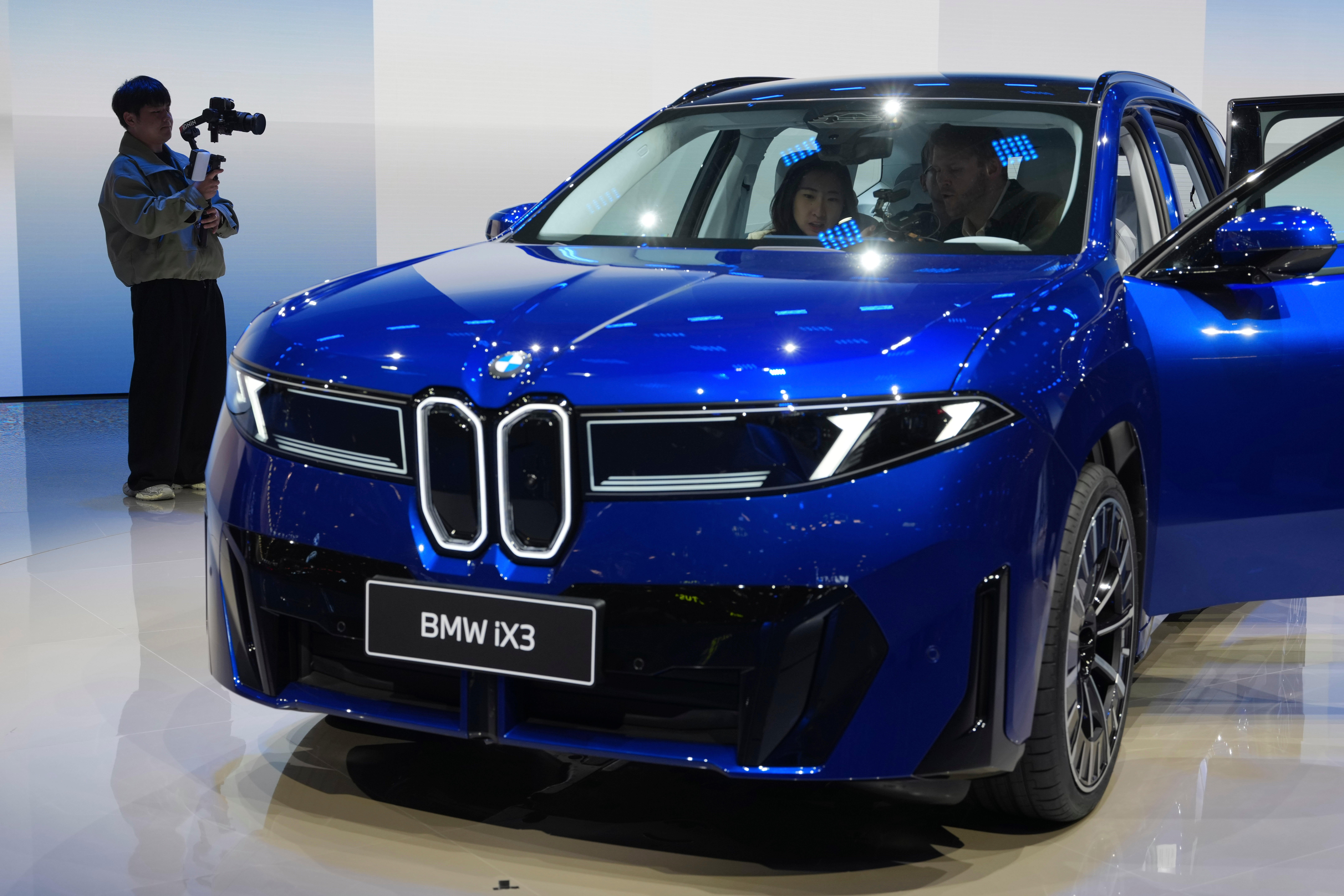 En blå BMW iX3 visas upp på en bilmässa. Två personer sitter i bilen och en person filmar den.