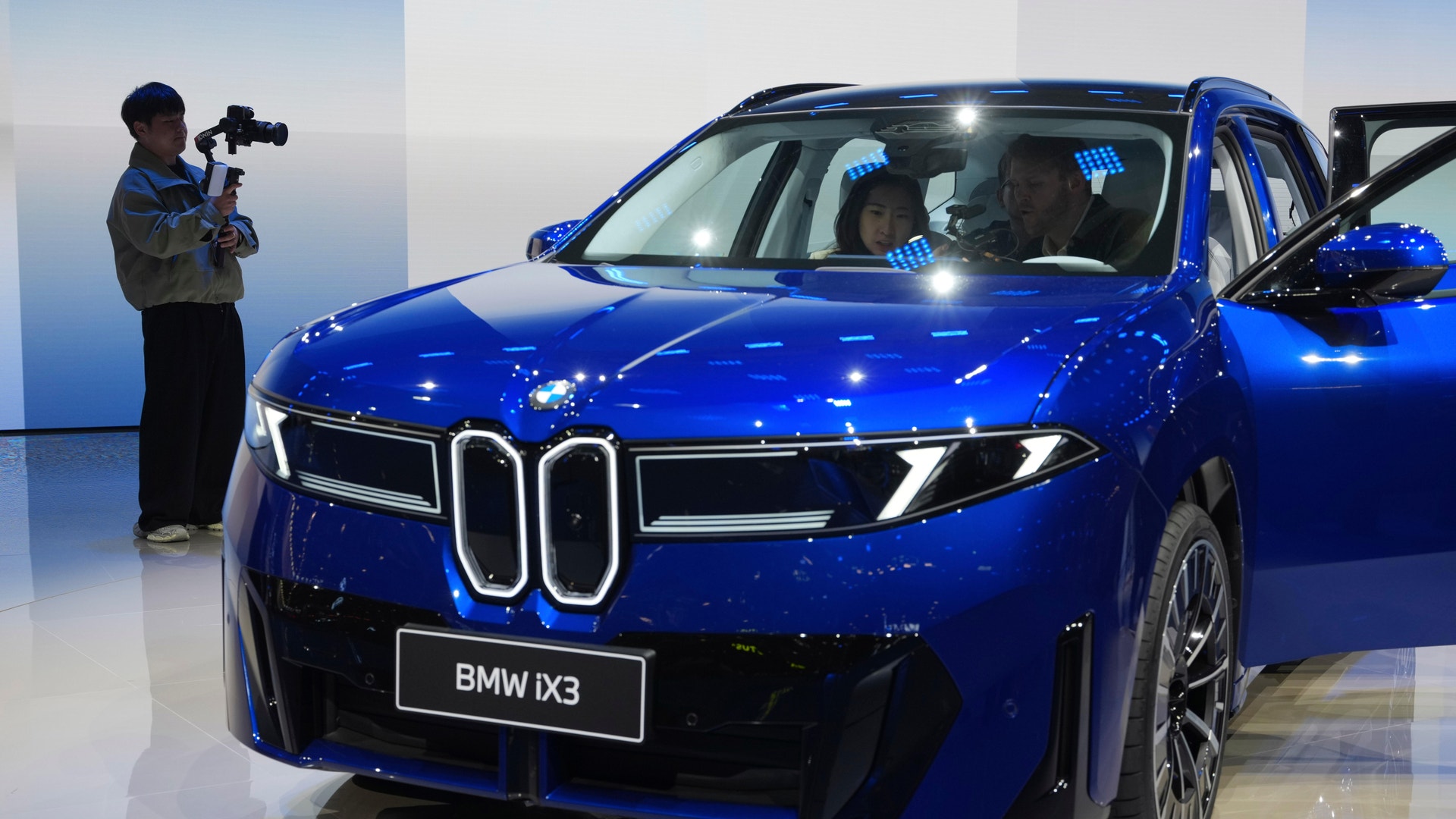 En blå BMW iX3 visas upp på en bilmässa. Två personer sitter i bilen och en person filmar den.