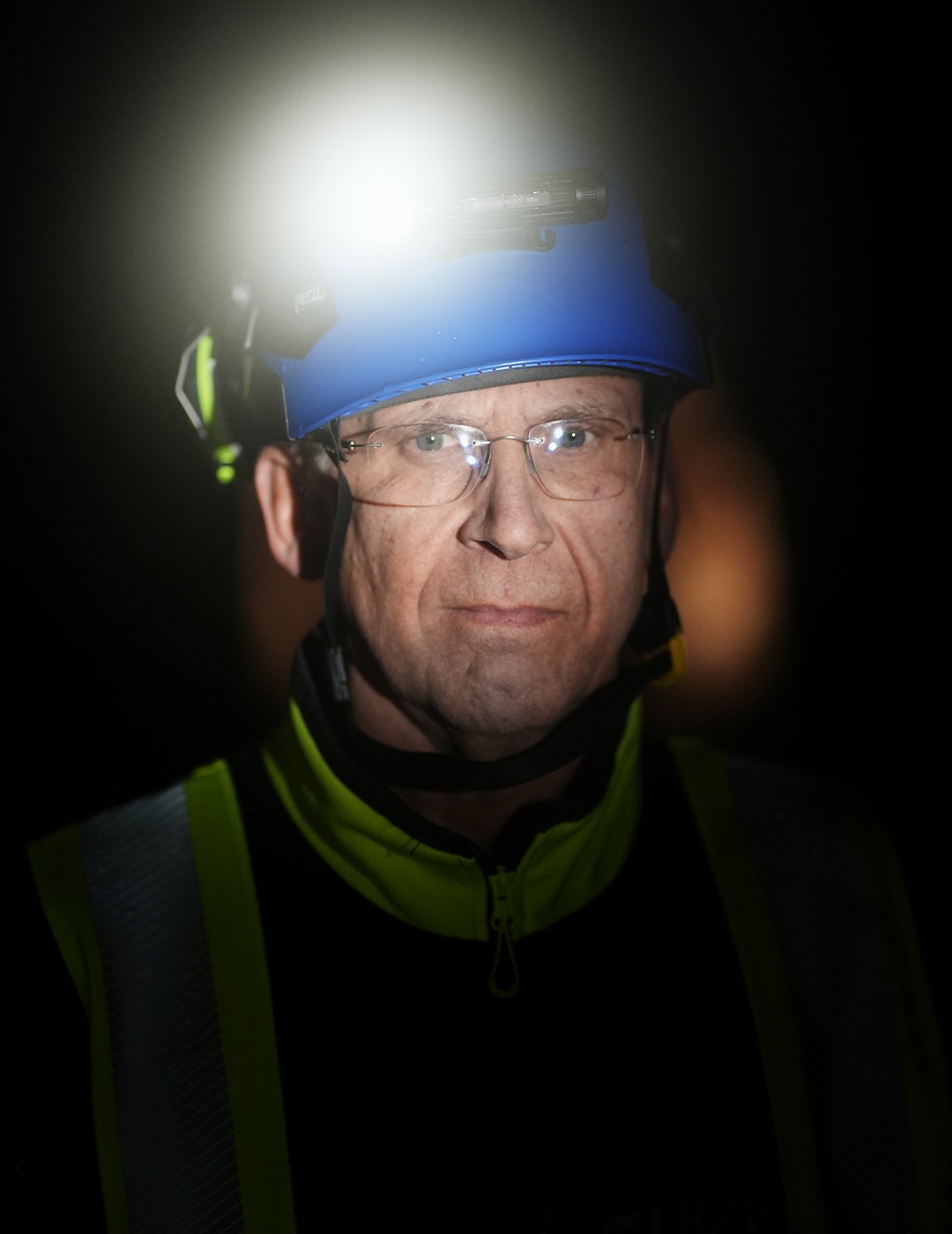 Porträtt av en man med blå arbetshjalm, glasögon och reflekterande väst med pannlampa