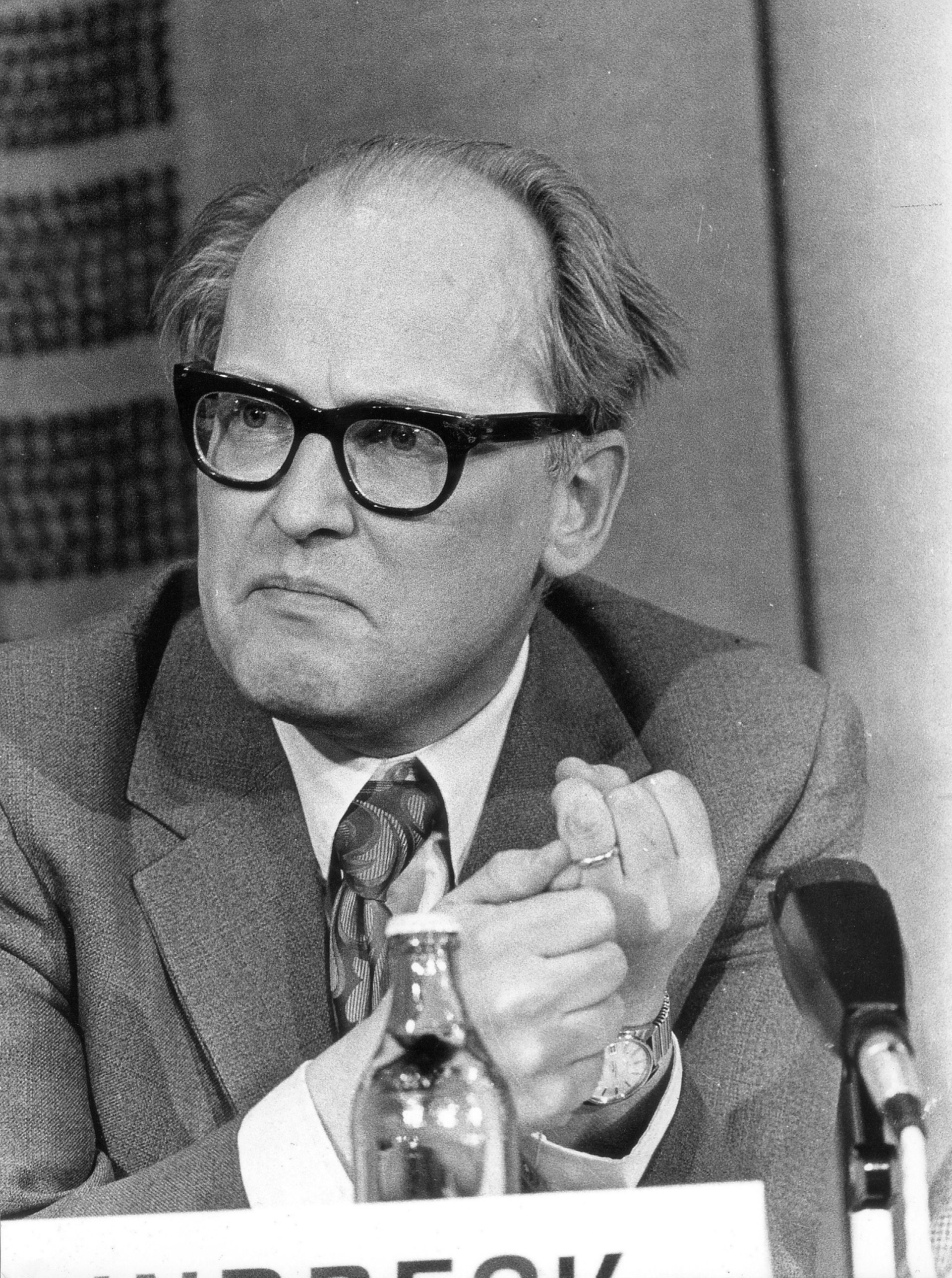 Svartvit fotografi av en äldre man med glasögon och kostym som sitter vid ett bord med mikrofon och pekar med fingret under vad som verkar vara en presskonferens.