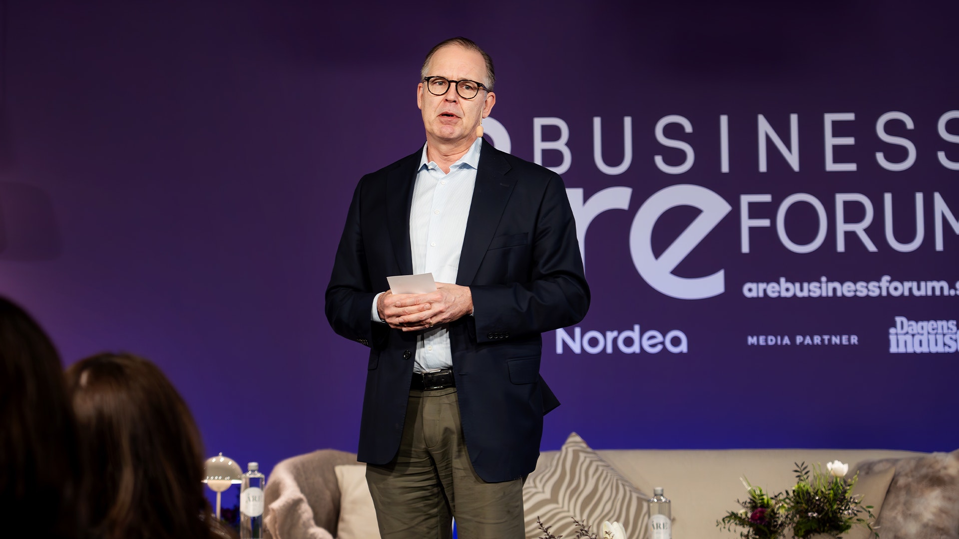 Man i kostym talar på en affärskongress framför en scen med lila bakgrund.