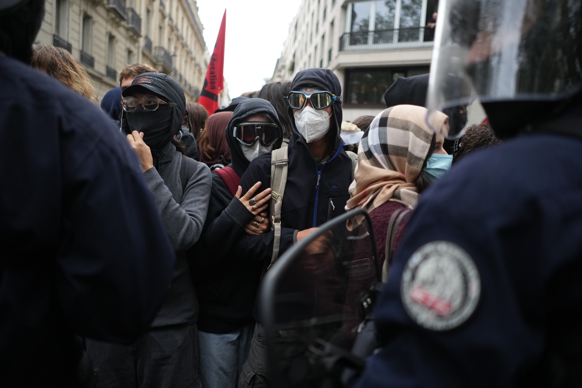 Maskerade demonstranter står tätt ihop på en gata framför poliser med sköldar.