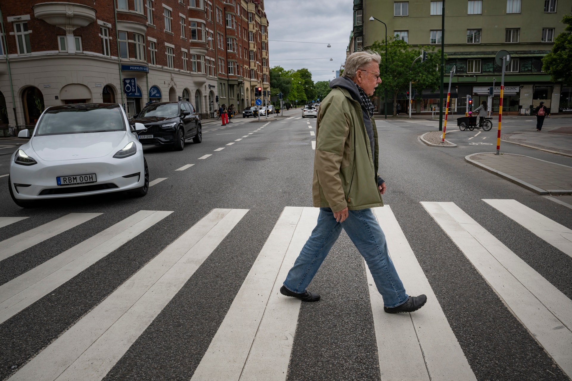 En äldre man går över ett övergångsställe på en stadsgata med bilar och byggnader i bakgrunden.