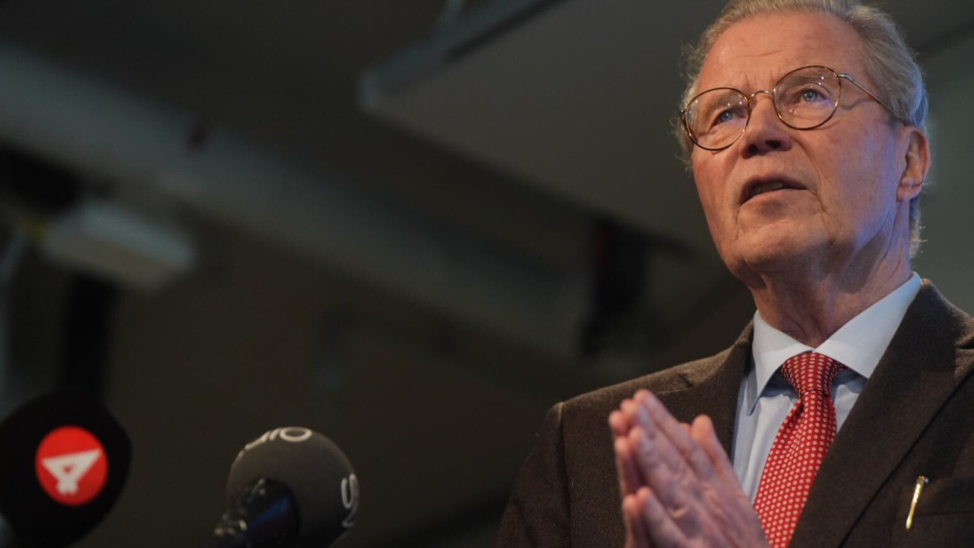 En äldre man i glasögon och kostym talar vid ett mikrofon under en presentation.