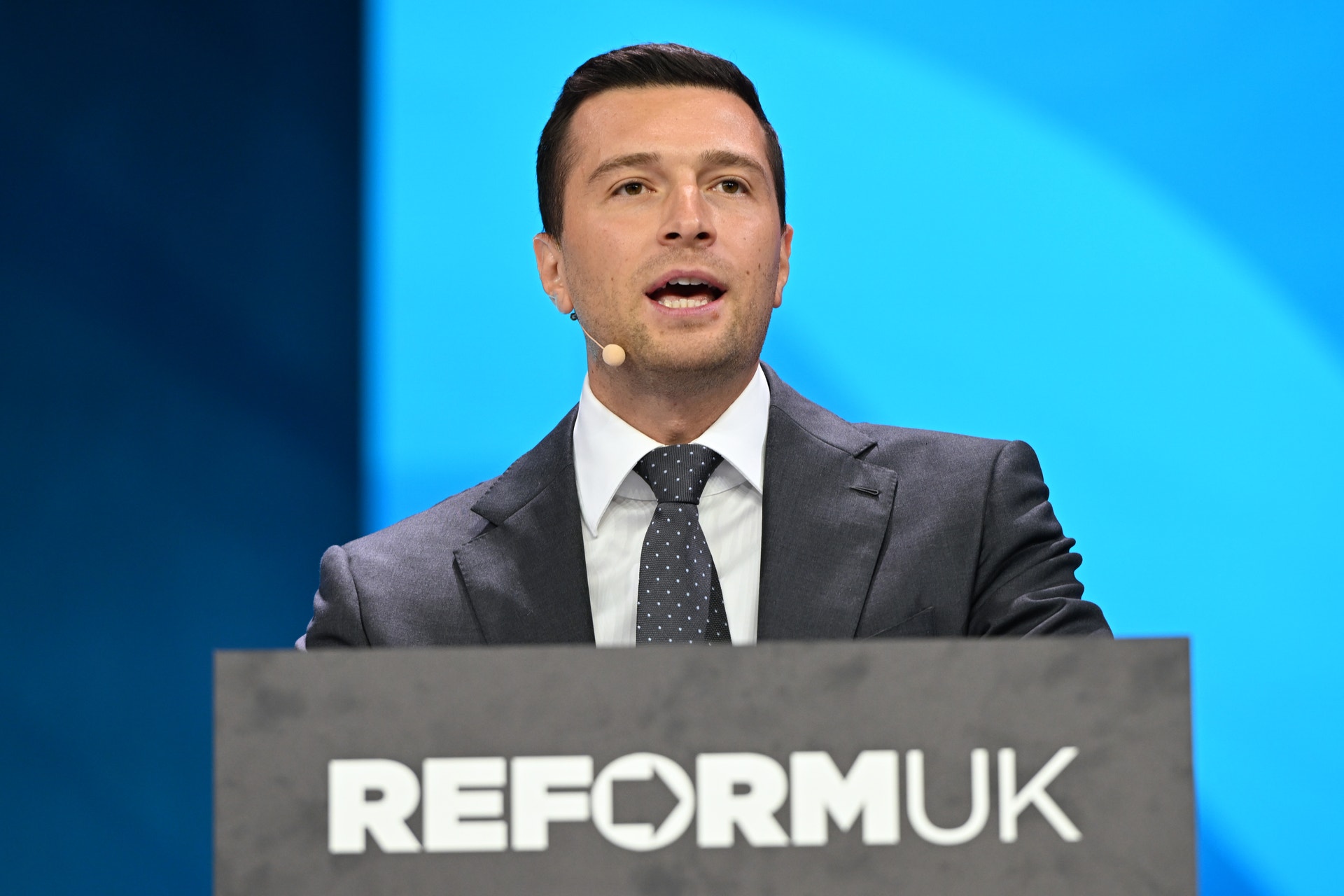 En man i kostym och slips talar i en headsetmikrofon bakom en talarstol med texten Reform UK.