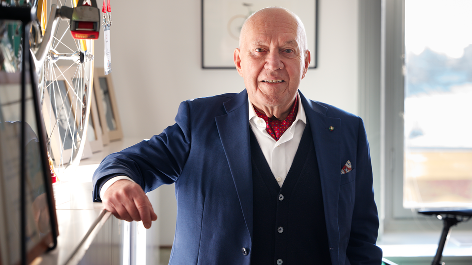 Porträtt av en äldre man i marinblå kavaj och röd slips som lutar sig mot en cykelhjul inomhus.