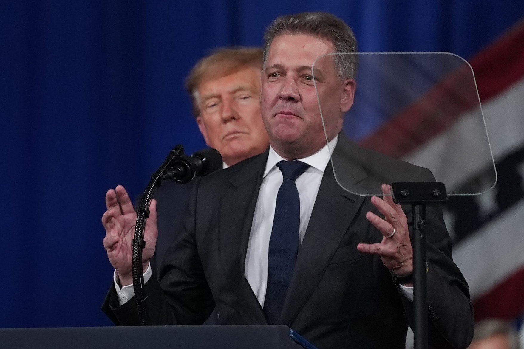 En man i mörk kostym och slips står vid en talarstol och gestikulerar med händerna. Bakom honom syns en annan man och en amerikansk flagga.