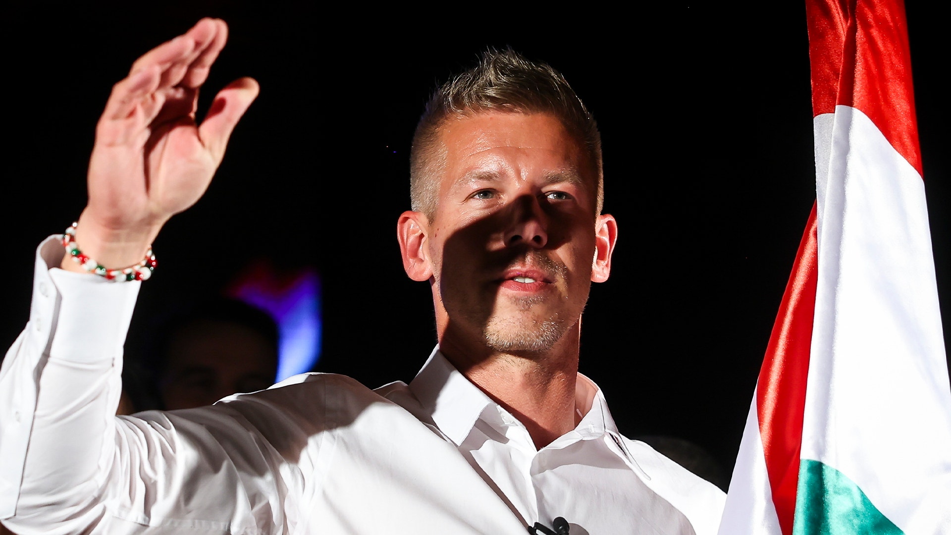 Man i vit skjorta vinkar mot publik med ungersk flagga i bakgrunden.