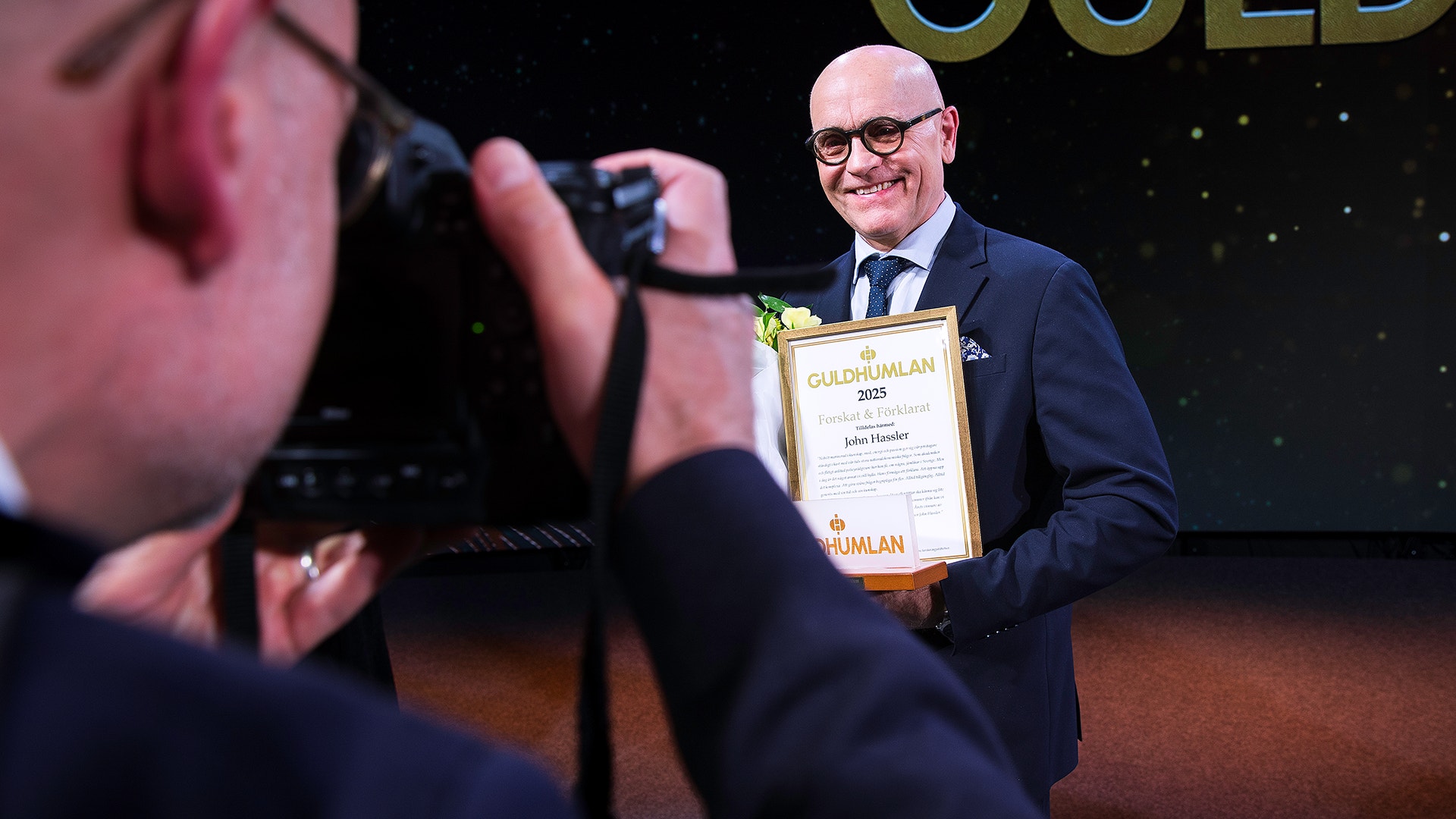 Man i kostym och glasögon håller ett diplom och poserar på en scen medan han fotograferas.