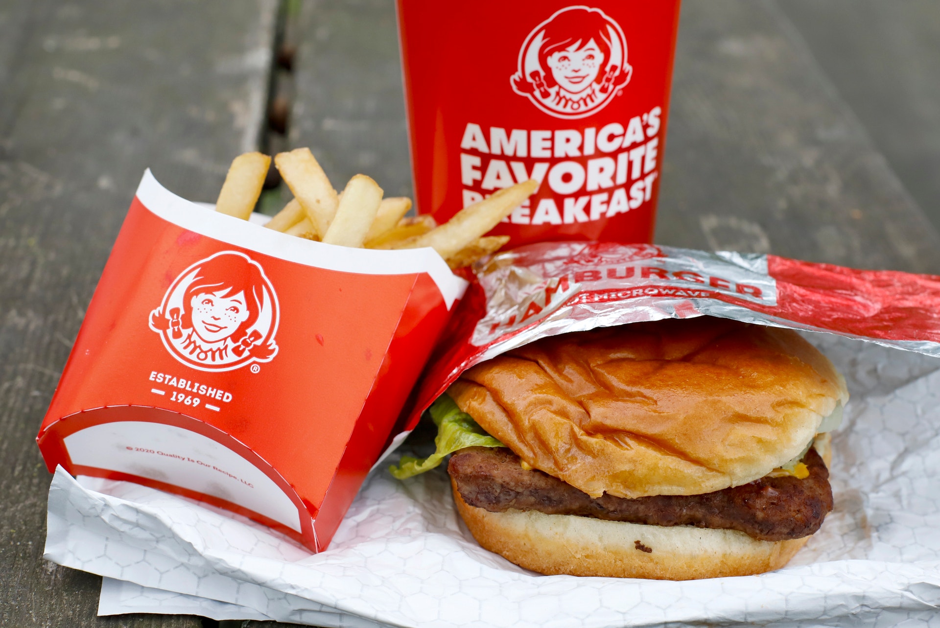 En hamburgare med ost och sallad, en röd ask med pommes frites och en röd dryckbägare med Wendys-logotyp på en grå yta.