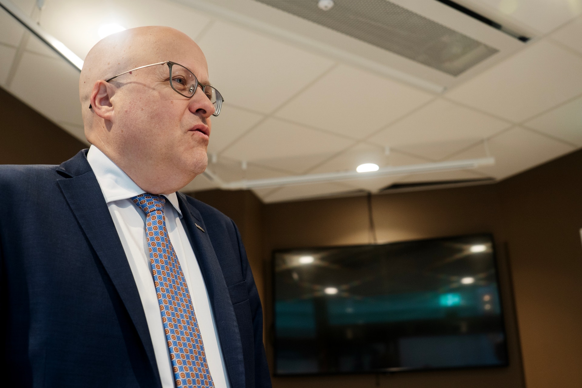 En man i glasögon och mörkblå kostym står i profil i ett möte eller konferensrum.