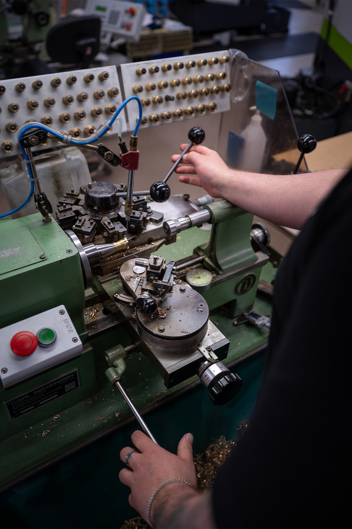En person arbetar med en grön industriell maskin. Personen håller i ett verktyg och utför reparation eller justering av maskinen. Maskinen har ett kontrollpanel med många små komponenter och knappar.