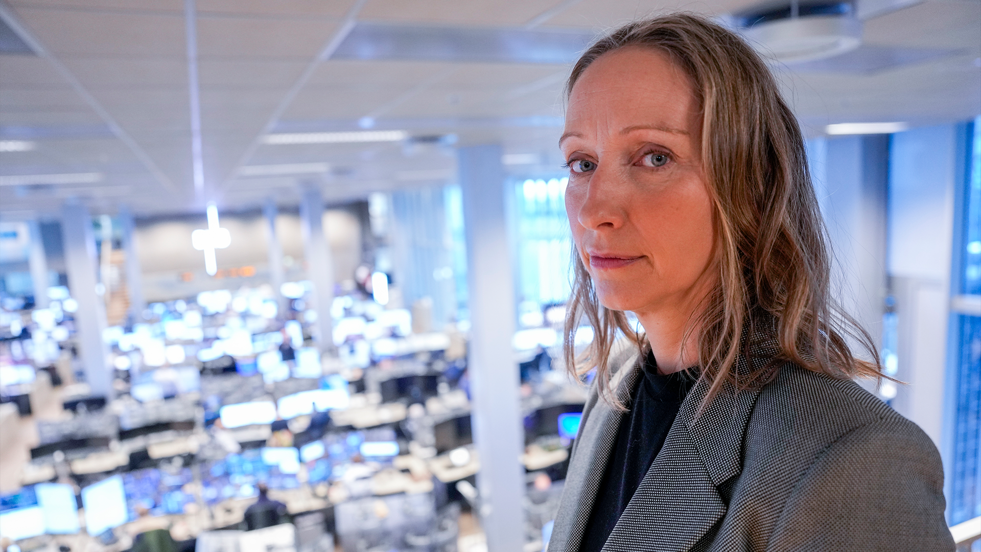 Porträtt av en kvinna i grå blazer som står i ett modernt kontorslandskap med många arbetsplatser i bakgrunden.