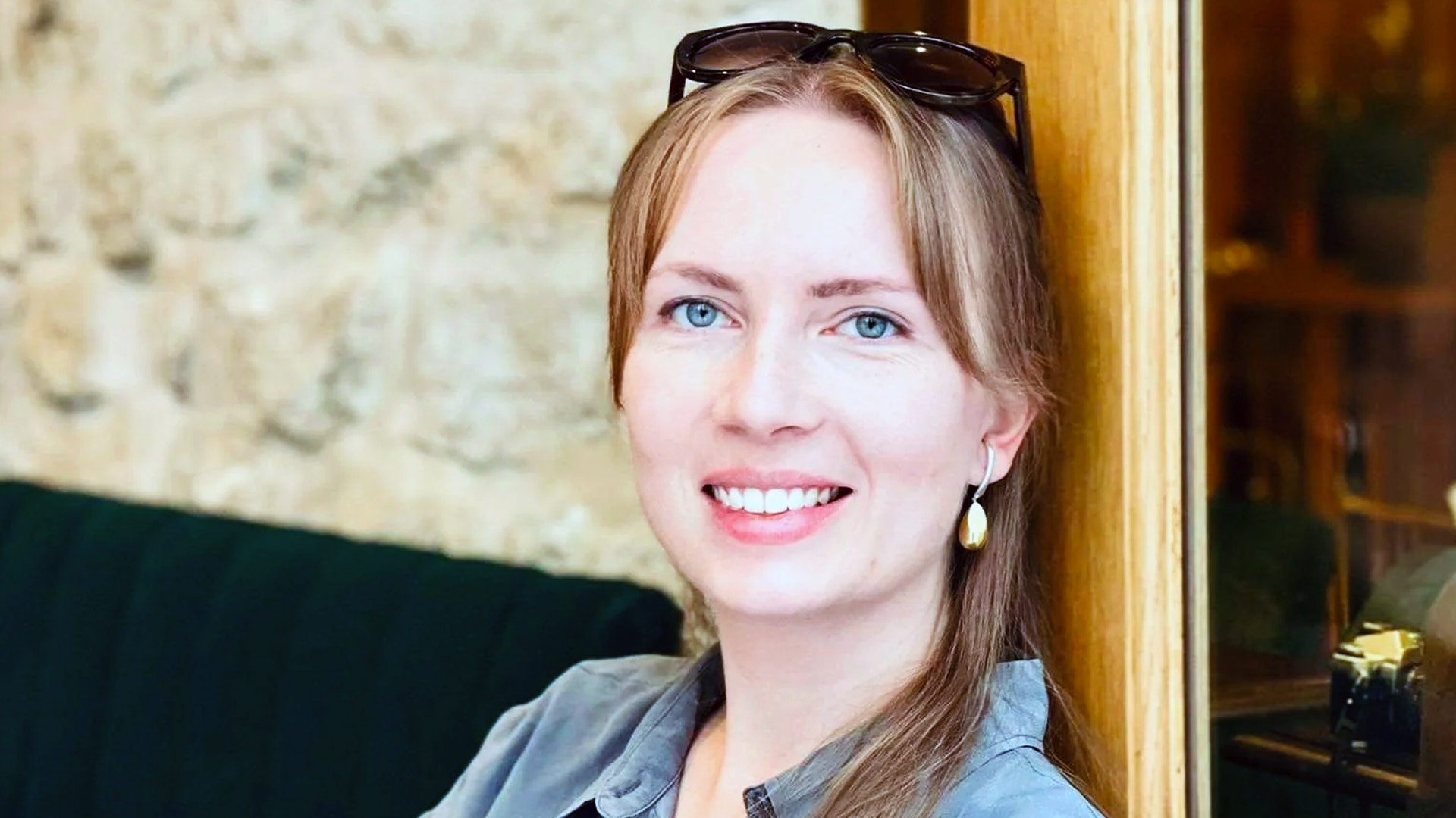 Porträtt av en leende kvinna med långt brunt hår och blå ögon. Hon bär en ljusblå skjorta och solglasögon på huvudet. Hon står framför en vägg med gul ram och ler mot kameran.