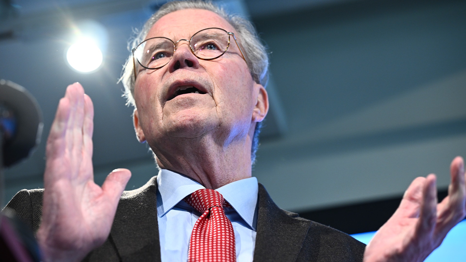 En man i glasögon och mörk kostym talar framför publik med höjda händer.