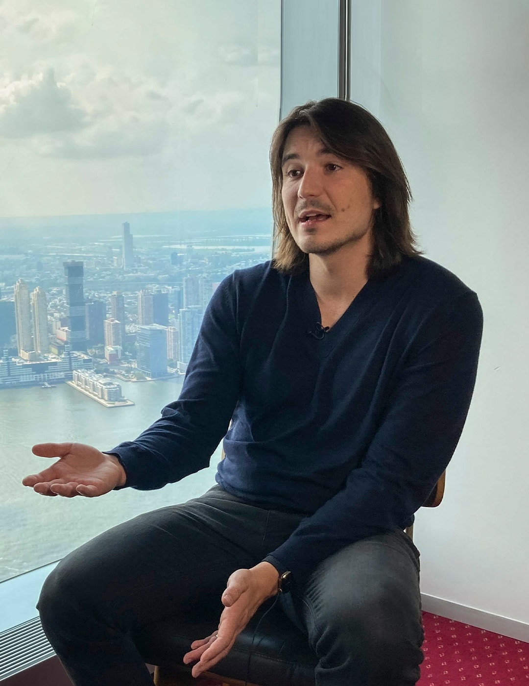 Man med långt mörkt hår sitter vid ett stort fönster med en stadsvy i bakgrunden. Han bär en mörkblå skjorta och gestikulerar medan han talar.