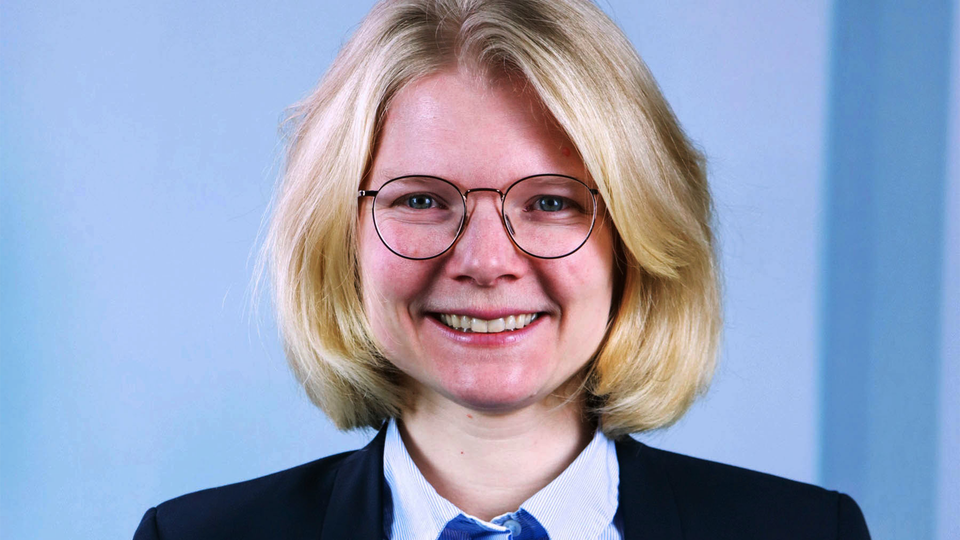 Porträttfoto av en person med blont hår och glasögon, klädd i mörkblå kavaj och vit skjorta mot blå bakgrund.