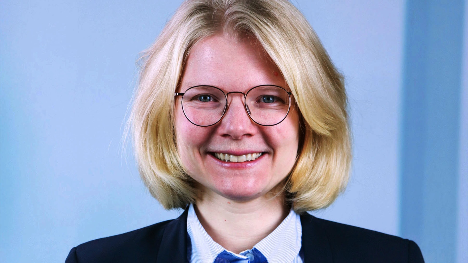 Porträttfoto av en person med blont hår och glasögon, klädd i mörkblå kavaj och vit skjorta mot blå bakgrund.