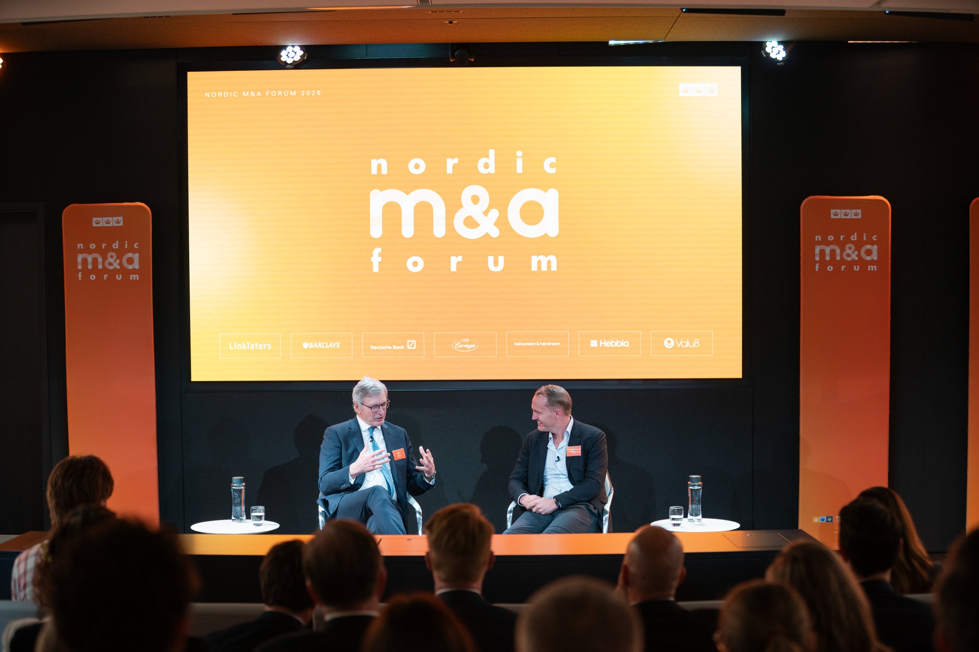 Två män på scen under en konferens diskuterar vid Nordic M&A Forum, med publik i förgrunden och en gul skärm bakom dem.